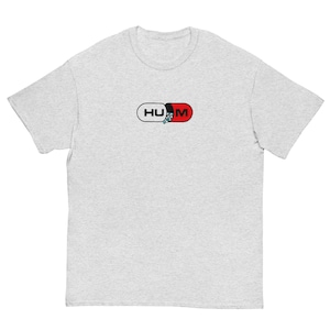 Könnte beinhalten: Ein hellgrau meliertes Kurzarm-T-Shirt mit einer Grafik auf der Brust, die eine in der Mitte geteilte Kapsel zeigt. Die linke Seite ist weiß mit den schwarzen Buchstaben HU, die rechte Seite ist rot mit dem schwarzen Buchstaben M und kleinen blauen Pillen.