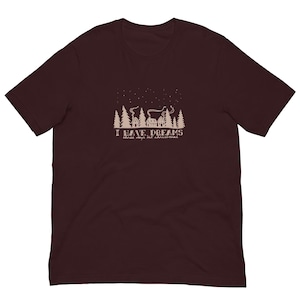 Può includere: T-shirt bordeaux con grafica color crema raffigurante una scena invernale con case, alberi e stelle. Il testo "I HAVE DREAMS three days til christmas" è stampato sotto.