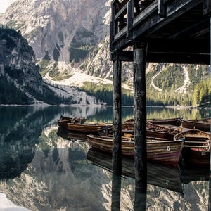 Può includere: Un molo di legno con diverse barche di legno ormeggiate. Le barche sono in un lago calmo con montagne sullo sfondo.