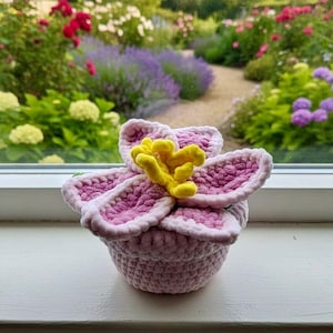 Könnte beinhalten: Eine handgefertigte Häkelblume in Rosa- und Gelbtönen. Die Blume hat große, geschichtete Blütenblätter und eine strukturierte Optik. Der Hintergrund zeigt einen Garten mit verschiedenen Blumen und Grün.
