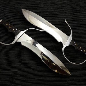 May include: Two polished steel swords with curved blades and black handles adorned with gold-colored studs. The swords have a distinctive design with a finger loop guard. The blades reflect the surrounding environment.