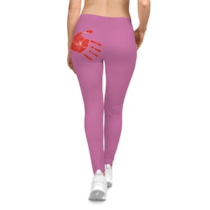 May include: Pink leggings with a red handprint design on the back. The leggings are form-fitting and made of a smooth material. The handprint is a bold, eye-catching graphic.