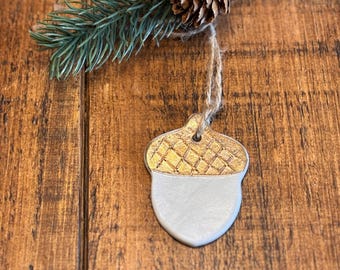 May include: An acorn-shaped decoration with a gold textured top and a white bottom, suspended by twine. A small pinecone and a sprig of evergreen are attached to the twine, set against a wooden background. Suitable for festive seasonal displays.