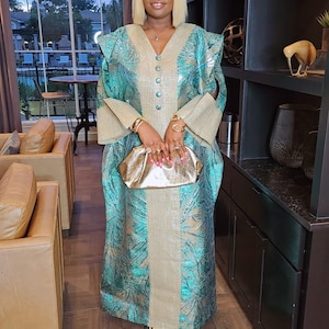 May include: A teal and gold patterned dress with a V-neck and button details. The dress has wide sleeves and is paired with a gold clutch. The outfit is completed with gold heels. The dress is a formal wear.