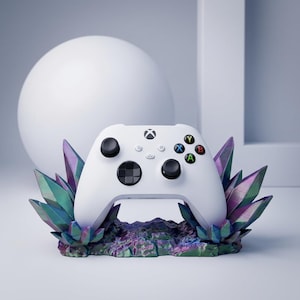 May include: A white Xbox controller rests on a decorative base with colorful, crystalline structures. The controller features black buttons and the iconic Xbox logo. The base has shades of purple, green, and blue.