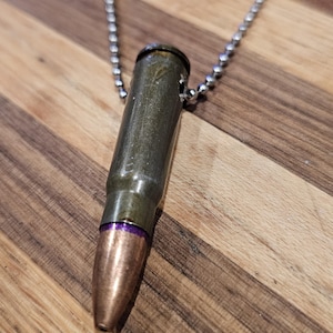 May include: A bullet-shaped pendant necklace. The pendant is a metallic olive green with a copper-colored tip and a purple band. It hangs from a silver ball chain. The necklace rests on a wooden surface.