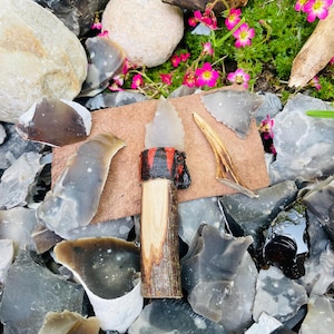 May include: A flint knife with a wooden handle wrapped in red and black leather. The knife is surrounded by flint stones and pink flowers.