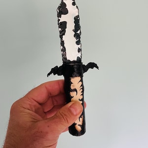 Könnte beinhalten: Ein handgefertigtes Messer mit schwarzem Griff und weiß-schwarzer Klinge. Der Griff weist ein beiges Blumendesign auf. Das Messer wird in einer Hand vor einem hellblauen Hintergrund gehalten.