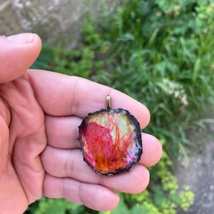 May include: A circular pendant with a gold bail, displaying an abstract design. The pendant's core features red, orange, and yellow colours, framed by a black border. The design has a natural, organic aesthetic.