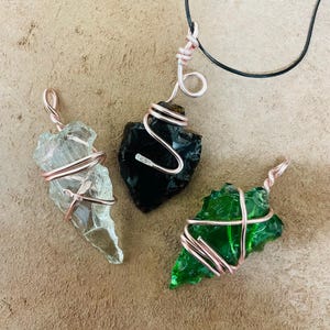 May include: Three wire-wrapped pendants featuring arrowhead-shaped glass. One is clear, one is black, and one is green. The pendants are suspended on a black cord. The wire is a copper color.