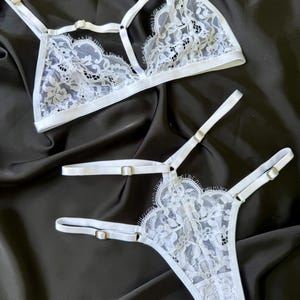Könnte beinhalten: Weißes Spitzen-Dessous-Set mit einem Triangel-BH und einem String-Tanga. Das Set hat verstellbare Träger und Metallbeschläge. Die Spitze hat ein Blumenmuster. Die Dessous werden auf schwarzem Hintergrund präsentiert.