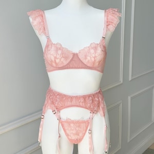 Könnte beinhalten: Ein blush-rosa Dessous-Set auf einer Schaufensterpuppe. Das Set besteht aus einem BH mit Spitzendetails und gerüschten Trägern, einem Strumpfhalter und einem String. Die Dessous sind aus zartem, transparentem Stoff gefertigt.