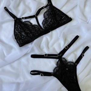 Puede incluir: Conjunto de lencería de encaje negro con un sujetador triangular con tirantes ajustables y un tanga a juego. El conjunto incluye delicados bordes festoneados y herrajes plateados. La lencería se muestra sobre un fondo de tela blanca.
