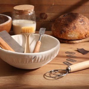 Op de afbeelding: Een broodmaakopstelling met een brood, een rijsmand en diverse gereedschappen. De gereedschappen omvatten een deegschraper, een borstel en een deeghark. Een pot met een kurken deksel en een kom met bloem zijn ook aanwezig.