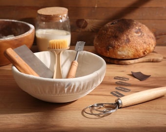 May include: A bread-making setup with a loaf of bread, a proofing basket, and various tools. The tools include a dough scraper, a brush, and a dough whisk. A jar with a cork lid and a bowl with flour are also present.