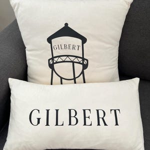 May include: Two off-white decorative throw pillows. One square pillow features a black graphic of a water tower with the word "GILBERT" printed on it. The other pillow is rectangular and has the word "GILBERT" printed in black.