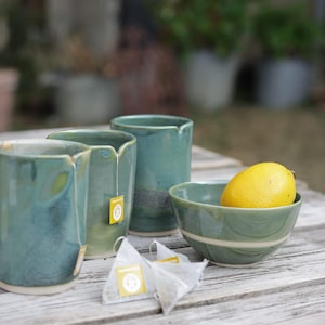 Peut inclure: Trois tasses cylindriques en céramique avec un émail bleu-vert, chacune présentant une petite encoche sur le bord. Un bol en céramique assorti contient un citron jaune. Deux sachets de thé pyramidaux blancs avec des étiquettes jaunes.