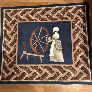 May include: A textile wall hanging featuring a woman using a spinning wheel. The woman is wearing a long dress and bonnet. The border has a repeating geometric design in brown, tan, and blue. The background is a dark blue. The overall design is reminiscent of a quilt.