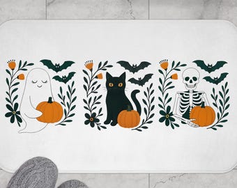 Puede incluir: Alfombra de baño rectangular blanca con temática de Halloween. El diseño presenta un fantasma, un gato negro y un esqueleto, cada uno sosteniendo una calabaza naranja. El diseño se completa con follaje verde, flores naranjas y murciélagos negros.
