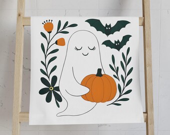 Könnte beinhalten: Weißes Handtuch zum Thema Halloween mit einem lächelnden Geist, der einen orangefarbenen Kürbis hält. Das Design umfasst dunkelgrüne Fledermäuse, Blumen und Blätter. Das Handtuch hängt an einer Holzleiter in einem Badezimmer.