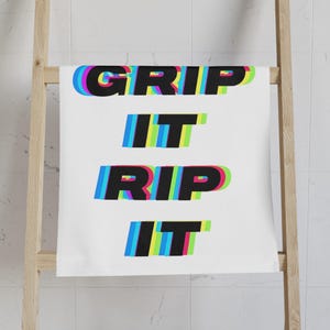 May include: White towel with the words "GRIP IT RIP IT" in a black, blue, green, and red layered font. The towel is hanging on a wooden rack in a bathroom setting.