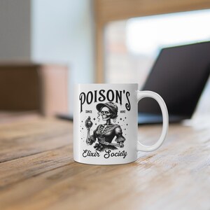 May include: White ceramic mug featuring a black and white illustration of a skeleton holding a potion bottle. The mug has the text "POISON'S" above the illustration and "Elixir Society" below. The mug is on a wooden surface.