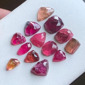 May include: A collection of faceted gemstones in shades of pink, red, and peach. The gemstones are displayed on a white surface. The shapes vary, including teardrop, oval, and irregular cuts. The gems have a polished, reflective surface.