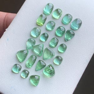Op de afbeelding: Een verzameling gepolijste, doorschijnende groene edelstenen in verschillende vormen, waaronder ovaal, druppelvormig en driehoekig. De edelstenen worden gepresenteerd op een wit oppervlak, waarbij hun helderheid en kleurvariaties worden benadrukt.