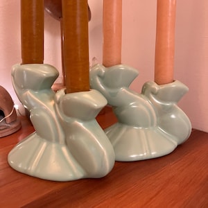 May include: A set of two teal ceramic candle holders shaped like stylized birds with three candle slots each. The holders are holding three candles, two brown and one peach.