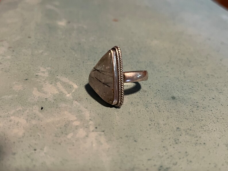 May include: A silver ring with a triangular shaped stone set in the center. The stone is clear with black lines running through it.