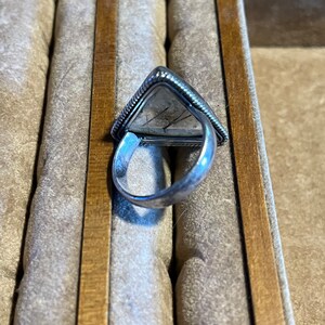 May include: A silver ring with a triangular shaped stone set in the center. The ring is in a box with a brown wooden edge.