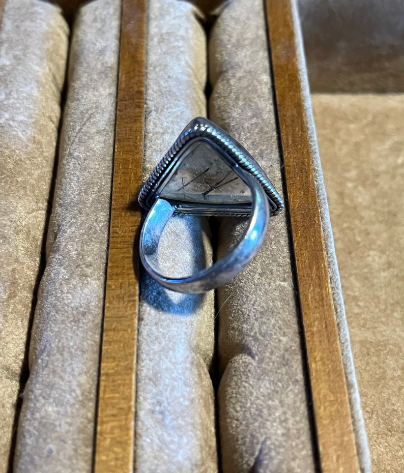 May include: A silver ring with a triangular shaped stone set in the center. The ring is in a box with a brown wooden edge.