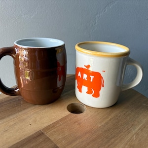 May include: Two ceramic mugs. One is brown with a light blue interior. The other is off-white with an orange illustration of an elephant and the word "ART". Both mugs have handles and are on a wooden surface.