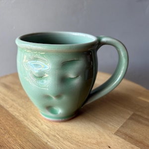 May include: A handmade ceramic mug in a soft green glaze. The mug features a sculpted face with closed eyes and a slightly open mouth. The handle is rounded, and the mug sits on a wooden surface.