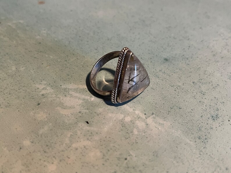 May include: A silver ring with a triangular clear quartz stone with black inclusions. The ring has a beaded edge.