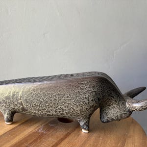 May include: A ceramic bull figurine featuring a textured, mottled dark gray and black glaze. The bull is depicted in a low-slung, horizontal stance with four short legs, a long body, and two prominent, curved horns extending from the head.