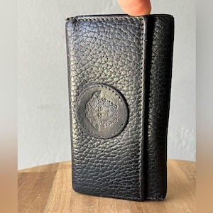 May include: Black leather key holder with a textured finish. The key holder features a round emblem with a detailed face design. The key holder is closed and has a vertical rectangular shape.