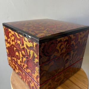 May include: A square, three-tiered stacking box featuring a glossy finish with a dense, intricate pattern of dark red flowers and leaves set against a textured, golden-yellow background. The lid and each tier have a smooth, black-edged rim.