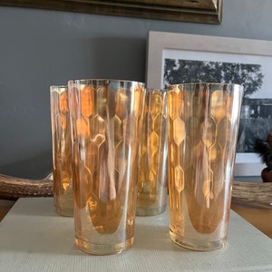 Puede incluir: Juego de cuatro vasos de vidrio ámbar con un patrón geométrico. Los vasos tienen un borde plateado y están dispuestos en fila sobre una superficie blanca.