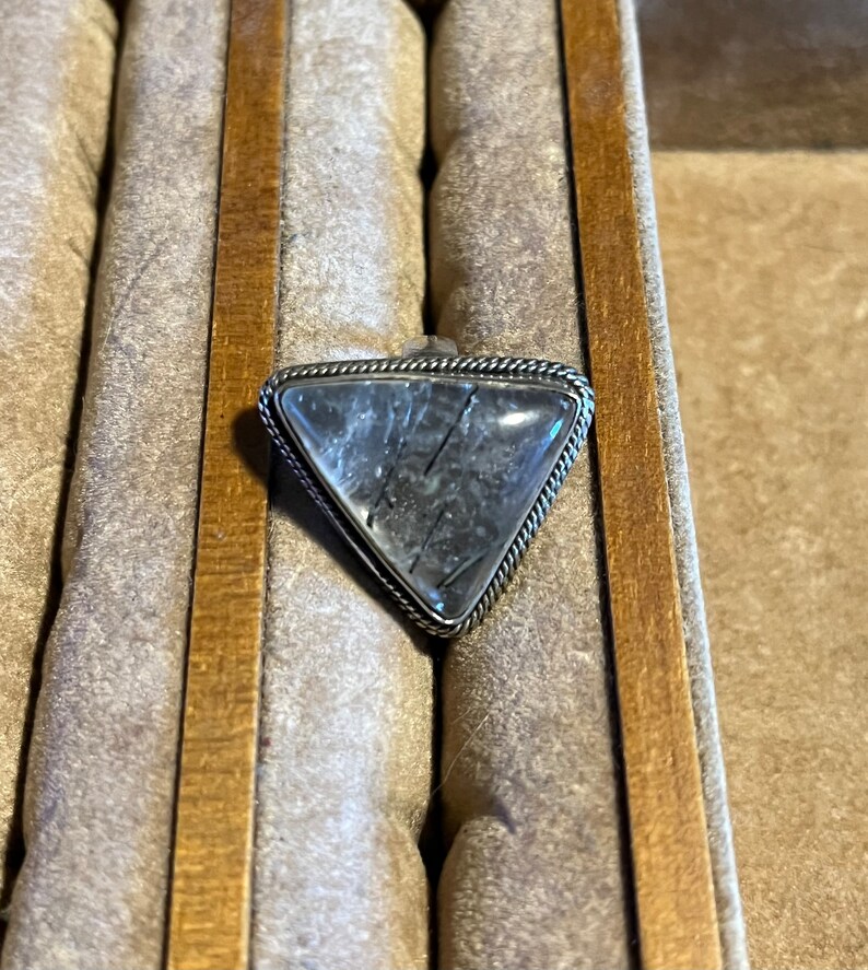 May include: A silver ring with a triangular clear quartz stone with black inclusions. The ring is in a wooden ring box.