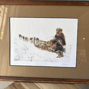 May include: Framed print titled "Homes of the North" depicting a person in a fur coat with a dog sled team on a snowy landscape. The artwork is in a brown wooden frame.