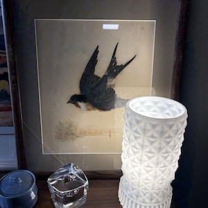 May include: A framed artwork featuring a black and white bird in flight, with a landscape background. A white, textured glass lamp and two small decorative objects sit on a wooden surface.
