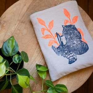 Op de afbeelding: Een lichtgrijze canvas tas met een tealgroene voering. De tas heeft een zeefdrukdesign van een wasbeer die een boek leest in een bossetting. De wasbeer is blauw en de bladeren zijn oranje.