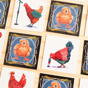 Könnte beinhalten: Eine Sammlung quadratischer Kunstdrucke mit skurrilen Illustrationen von Hühnern und Küken. Die Kunstwerke zeigen ein Küken in einem blauen Rahmen, ein Huhn mit Mikrofon und andere farbenfrohe Designs zum Thema Huhn. Die Drucke sind auf einer hellen Holzoberfläche angeordnet.