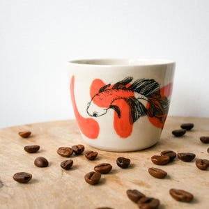 Peut inclure: Une tasse en céramique de couleur crème ornée d'une illustration à l'encre noire représentant un poisson combattant aux nageoires fluides, agrémentée de formes abstraites peintes en orange vif. La tasse est sans anse et possède une finition lisse.