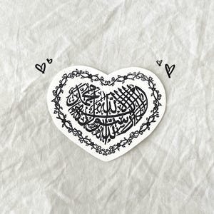 May include: A black and white sticker of a heart with a decorative border. The heart contains Arabic calligraphy.