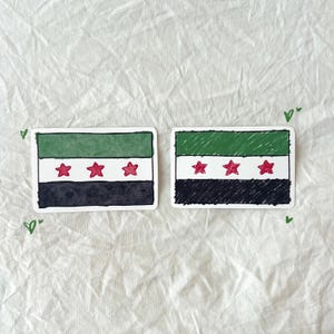 May include: Two stickers featuring the flag of Syria. Each sticker has a green stripe at the top, a white stripe in the middle, and a black stripe at the bottom. Three red stars are in the white stripe.