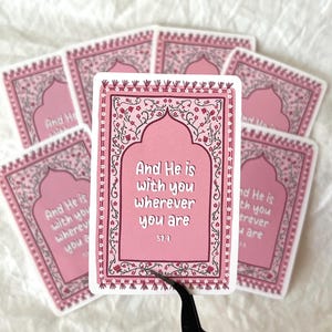 May include: Pink and white sticker with an ornate border and a quote from the Quran: "And He is with you wherever you are."  The quote is in a pink archway with a floral design. The sticker is 57:4.