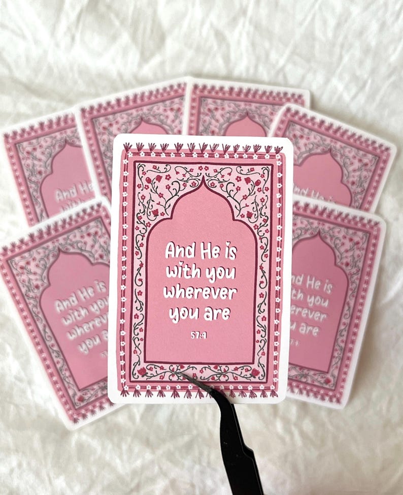 May include: Pink and white sticker with an ornate border and a quote from the Quran: "And He is with you wherever you are."  The quote is in a pink archway with a floral design. The sticker is 57:4.