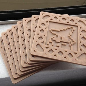 May include: A stack of six square, tan-colored coasters. Each coaster features a pixelated image of a creature within a decorative border. The coasters are made of a solid material and are stacked at a slight angle.